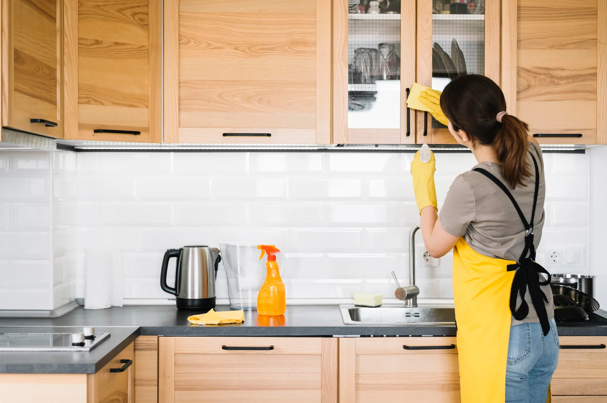 A woman cleaning kitchen cabinets with a yellow cloth and cleaning supplies