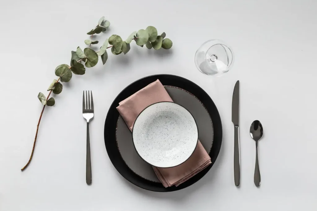Elegant minimalist table setting featuring black plates speckled ceramic bowl and soft pink napkin complemented by a single eucalyptus branch Elegant minimalist table setting featuring black plates speckled ceramic bowl and soft pink napkin complemented by a single eucalyptus branch