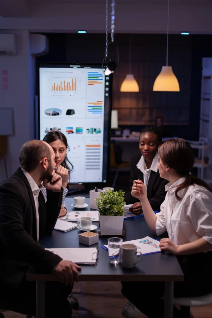 A diverse group of professionals in a meeting room discussing data displayed on a large screen with charts and graphs