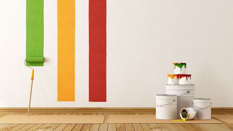 Una parete parzialmente dipinta con strisce verdi gialle e rosse accanto a una pila di barattoli di vernice bianca con gocce colorate e un rullo di vernice appoggiato al muro su un pavimento di legno