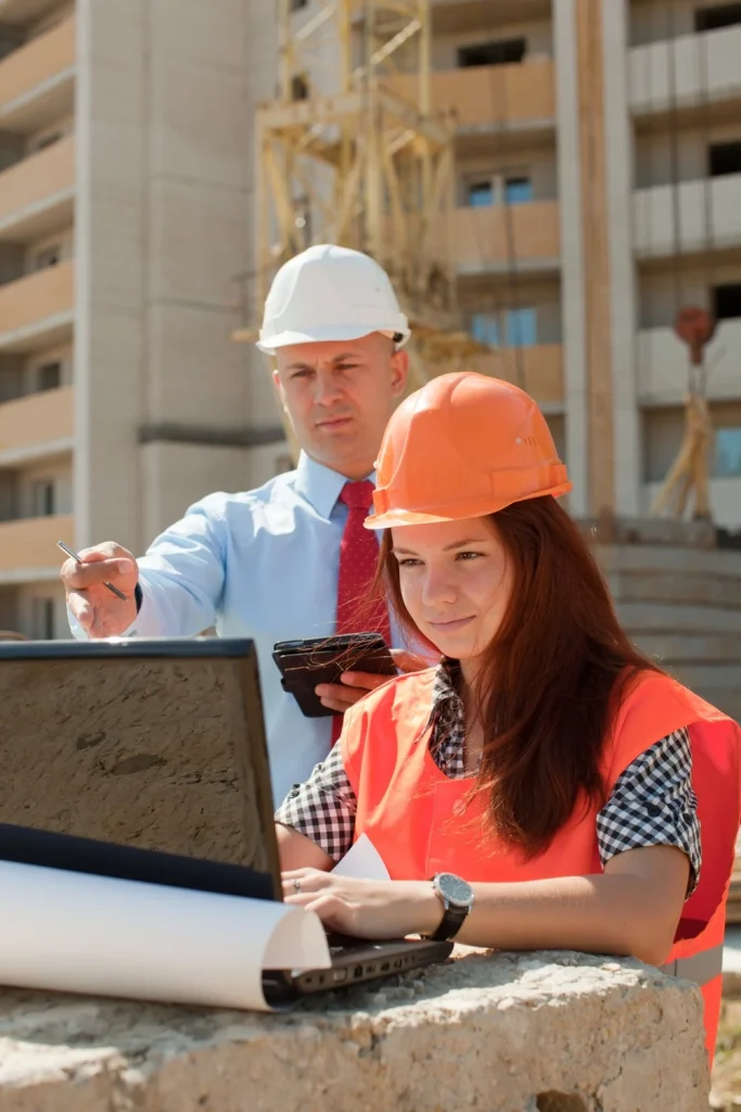 Construction workers in orange vests and helmets reviewing plans on a laptop at a construction site with one pointing at the screen