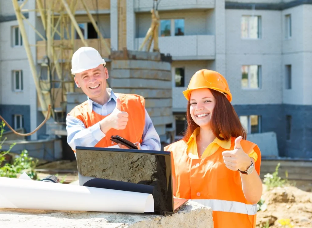 Two construction workers in orange vests and helmets giving a thumbs up at a construction site with a laptop and blueprints in front of them