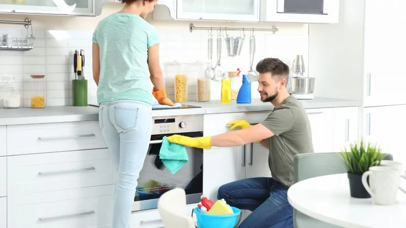Couple cleaning the kitchen together with one wiping the oven and the other cleaning the countertop